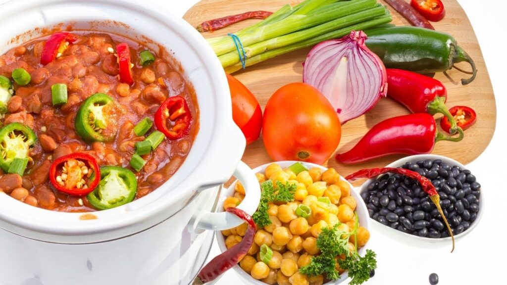 slow cooker with a bean stew on boards surrounded by colourful vegetables on a chopping board